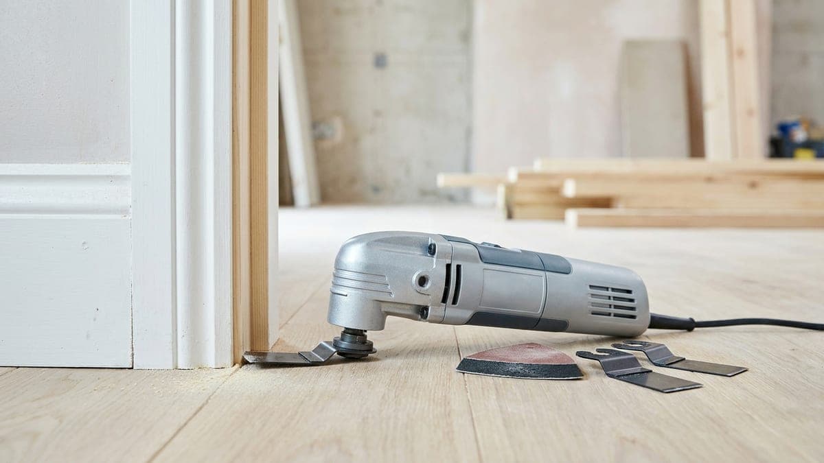 Outil multifonction oscillant en coupe plongeante dans une plinthe en bois, lame segment au ras du sol et sciure fine au premier plan